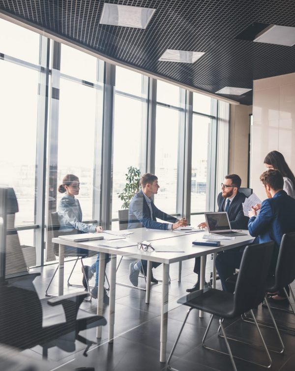 Blick in ein Geschäftsmeetingzimmer, in dem das Team von 5 Leuten (2 Frauen, 3 Männer) in einer Besprechung ist. Papierdokumente und Laptops sind auf dem Tisch.